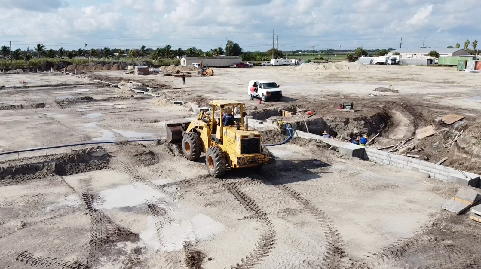 Aerial view of the Block Forever site in Moore Haven, FL — a wheel loader grading the cleared lot with foundation work and CMU block walls in progress
